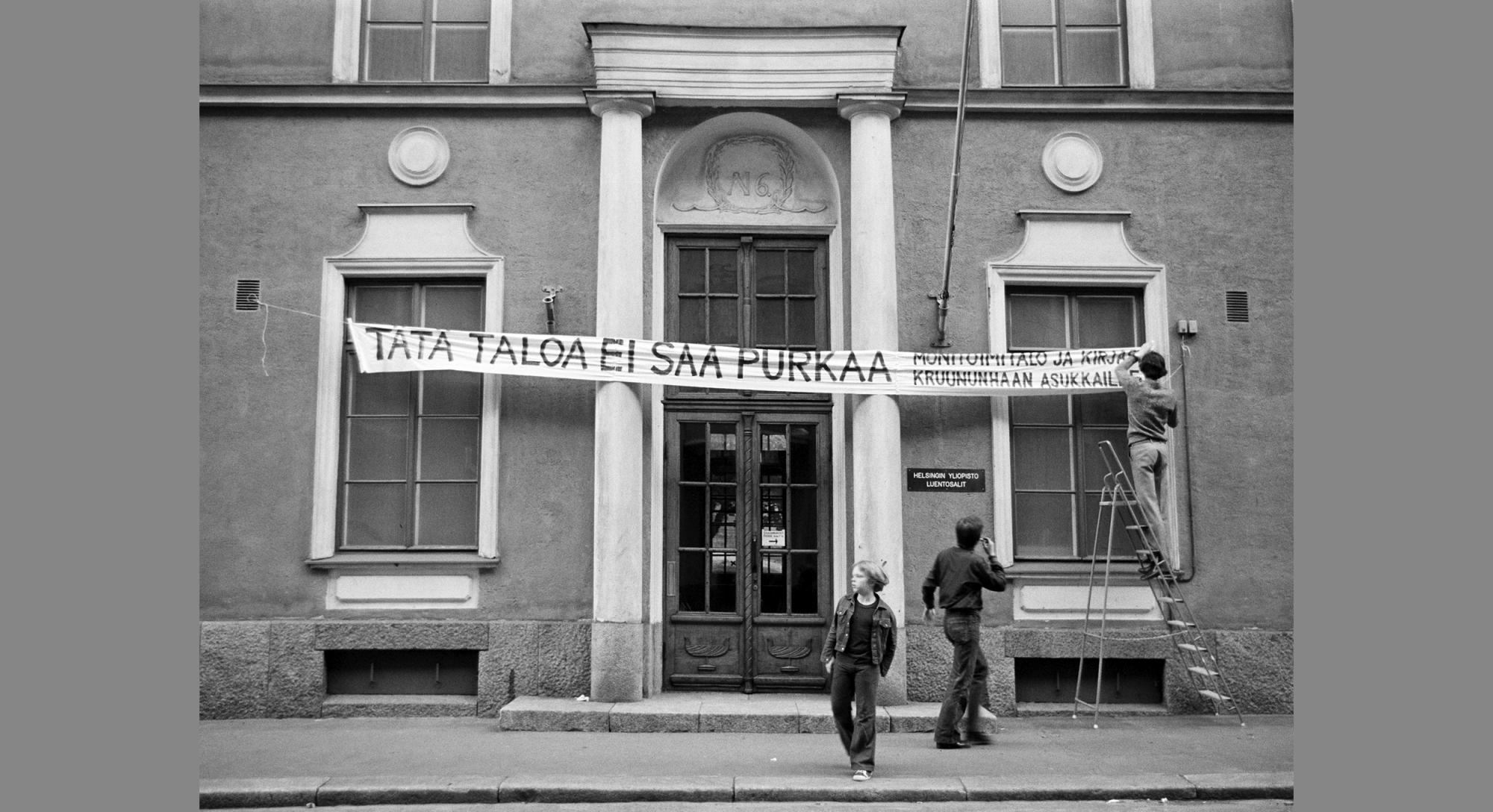 Suuren rakennuksen kadunpuoleiset ovet ja oven yllä itsetehty banderolli, jossa lukee "Tätä taloa ei saa purkaa. Monitoimitila ja kirjasto Kruununhaan asukkaille."