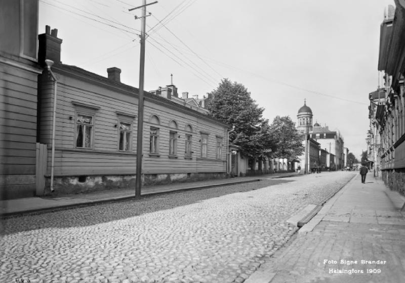 Mukulakivikatu, jonka reunalla pitkähkö puinen rakennus ja taustalla pilkottaa valkoisen kirkon torni.